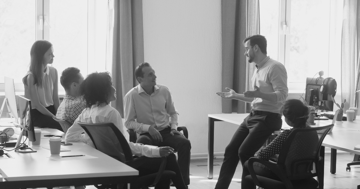 An office scene with a sales team lead and colleagues at desks, engaged in discussions to build a high-performing team.