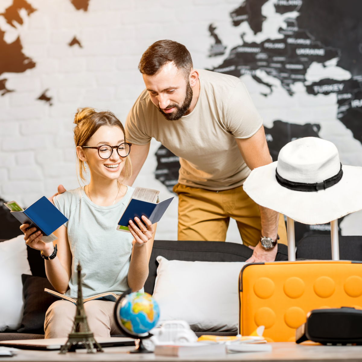 A man and woman sit on a couch with a suitcase and a hat, suggesting they are preparing for a trip or vacation.